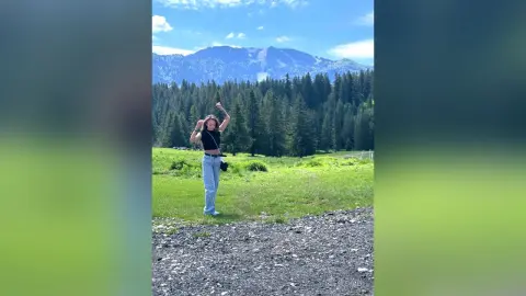 LesleyRed stands in a green field with mountains and trees in the background, wearing a black crop top and light blue jeans.
