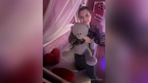 Young woman in a zebra print top hugging a grey teddy bear in a cozy bedroom setting.
