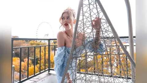 DaisyLin poses behind a silver mesh hanging chair, wearing a light blue off-the-shoulder lace top with her blonde hair styled up.