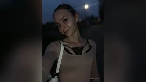 Smiling young woman in a beige long-sleeve top and black lace camisole posing outdoors at night.