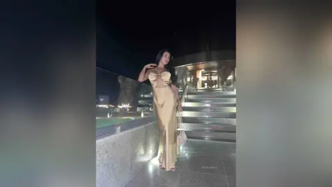 NicoleBella stands on outdoor stone steps wearing a long, tight, tan-colored cut-out dress.