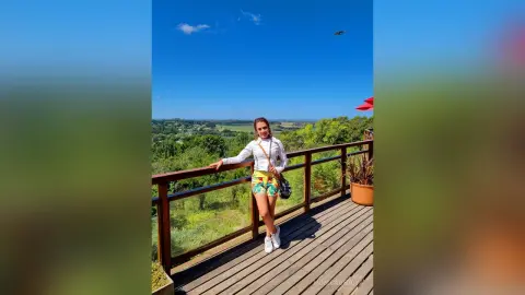 Young woman in white top and floral shorts relaxing on a sunny wooden balcony overlooking a lush green valley.