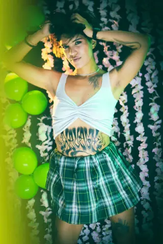 Young woman in a white tank top and plaid skirt posing against a floral wall with green lighting.