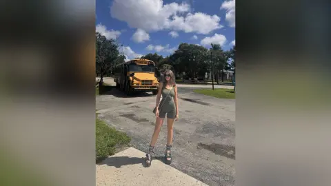 EvelynMills stands on a paved path wearing an olive green bodysuit and strappy sandals with a yellow school bus behind her.