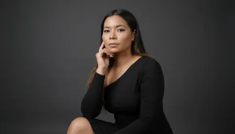 XinemaBonet poses against a dark grey background, wearing a long-sleeved black dress with her hand resting against her face.