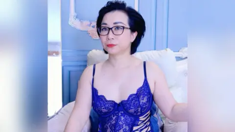 Woman with short dark hair wearing a blue lace bodysuit sitting on a white bed in a bright blue room.