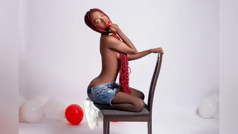 Red-haired woman in denim shorts sitting on a chair in a bright studio setting with red and white balloons.