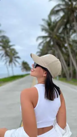 Smiling woman in white tank top and beige cap walking along a tropical palm tree path on a cloudy day.