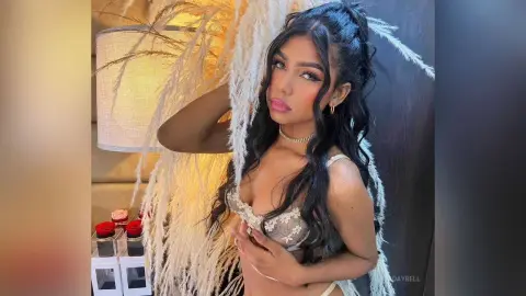 SadayBell poses in a lacy white bra, showing her long dark hair and gold choker against a background of white feathery decor.