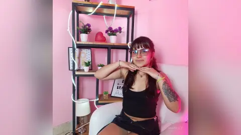 Young woman in a black tank top sitting in a pink room next to a shelf with flowers and fairy lights.