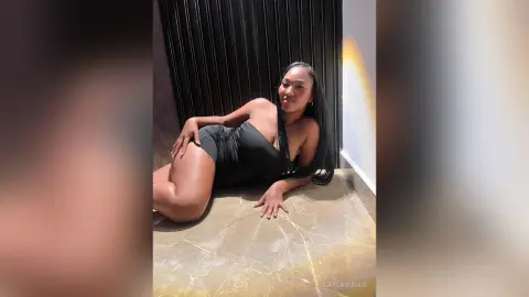 Confident woman in a sleek black bodysuit lounging on a marble floor against a dark slatted wall.
