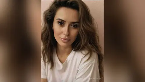 Portrait of a woman with wavy brown hair wearing a white t-shirt in a soft, natural light setting.