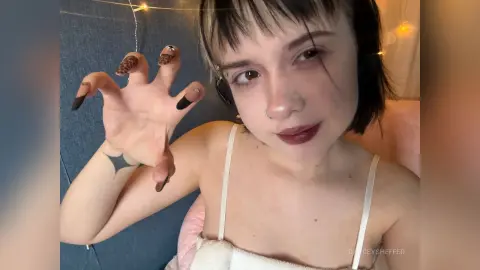 Young woman with short dark hair and nose ring wearing a white camisole in a cozy indoor setting.