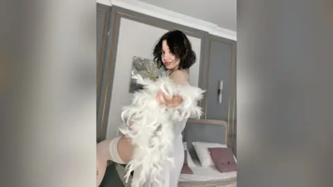 Elegant woman in a white feathered gown posing in a modern, bright bedroom setting.