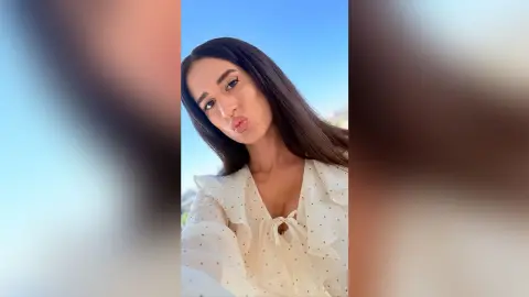 BellaLeen pouts her lips while wearing a white polka-dot blouse with ruffles and a tie-front, set against a clear blue sky.