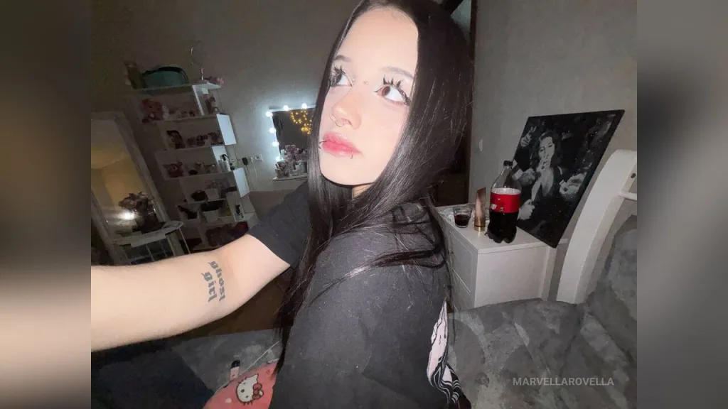 Young woman in a black long-sleeve top sitting in a dimly lit modern bedroom with a moody atmosphere.