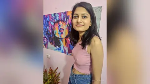 Young woman in a pink tank top smiling in front of colorful abstract wall art in a bright room.