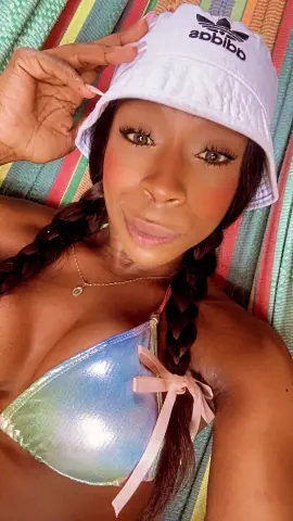 Smiling Black woman in a white bucket hat and iridescent bikini relaxing on a colorful striped beach towel.