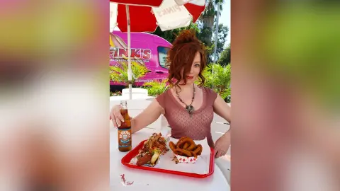 Redheaded woman in a pink top enjoying hot dogs and fries at a sunny outdoor food truck stand.