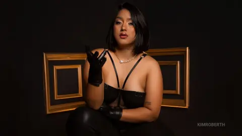 KimRoberth poses in black leather lingerie and gloves against a dark background with gold-trimmed frames.