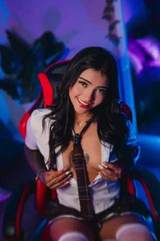 Smiling woman in a white shirt and tie sitting in a gaming chair with moody blue lighting.