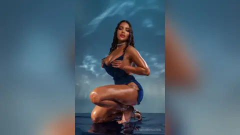 CassandraDuque poses kneeling in a blue cutout bodysuit, showing off her tanned skin and long dark hair against a cloudy blue background.