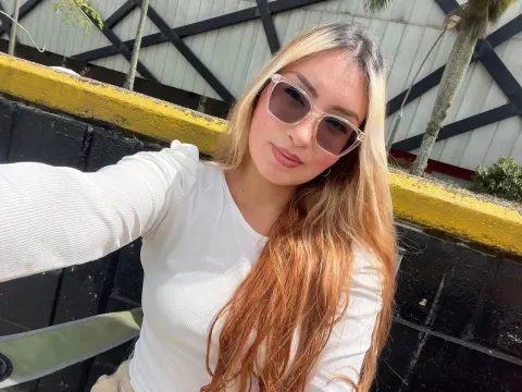 Ninablaze27 wearing sunglasses and a white long-sleeve shirt, posing outdoors with her long blonde and reddish hair.