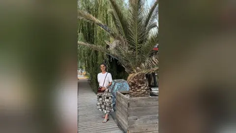 Smiling woman in a white blouse and patterned skirt standing by palm trees on a wooden boardwalk.