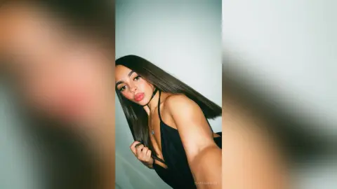 Young woman with long dark hair wearing a black halter top against a minimalist white wall background.
