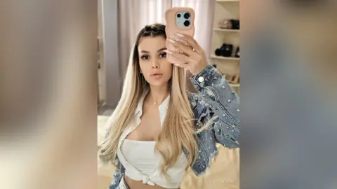 AshPayton poses for a mirror selfie wearing a white top and a studded denim jacket, showing off her long blonde hair.