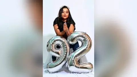 Happy woman in black outfit celebrating 92nd birthday with large silver foil balloons against white background.