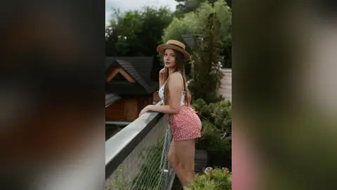 AdelinaKael poses on a balcony wearing a white tank top, red checkered shorts, and a tan sun hat, looking back over her shoulder.