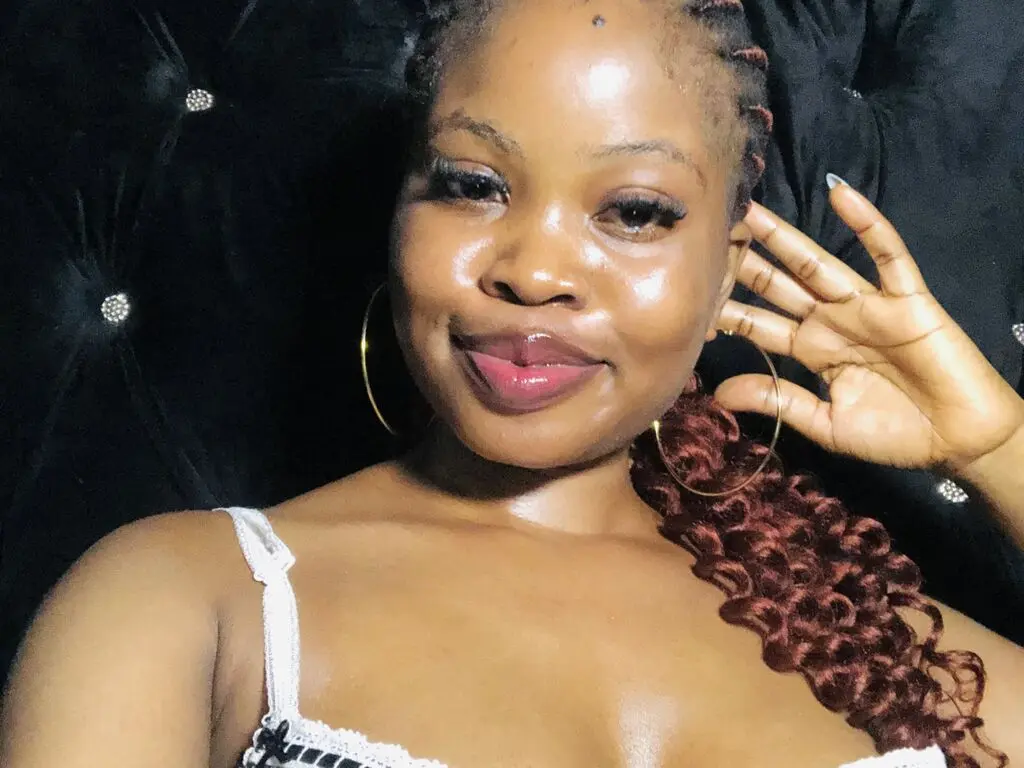 Smiling Black woman with long braids and hoop earrings wearing a white lace top in a cozy dark setting.
