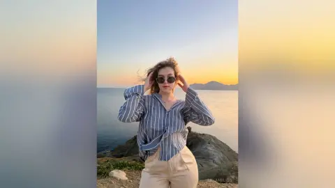 Young woman in a blue striped shirt and beige trousers posing by the ocean during a golden sunset.