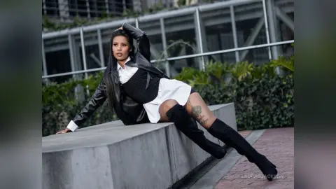 Stylish woman in a black leather jacket and white shirt leaning on a concrete ledge in an urban setting.