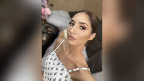 Elegant woman in a white floral patterned top with glamorous makeup posing in a soft-lit bedroom setting.