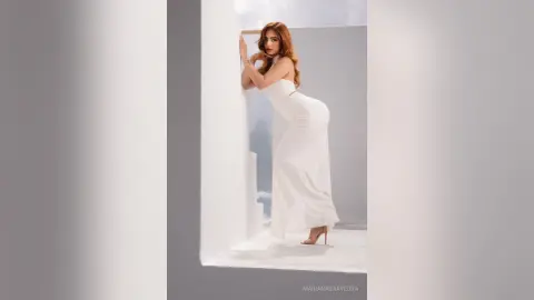 Elegant woman in a white evening gown posing gracefully in a bright, minimalist studio setting.