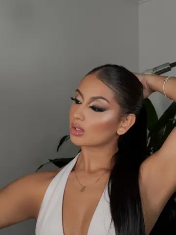 Glamorous woman with sleek high ponytail and smoky eye makeup wearing a white halter neck top indoors.