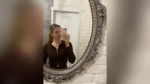 Young woman in a brown long-sleeve top taking a mirror selfie in a bright, minimalist room.