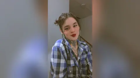 Smiling young woman with tattoos wearing a blue plaid flannel shirt in a cozy indoor setting.