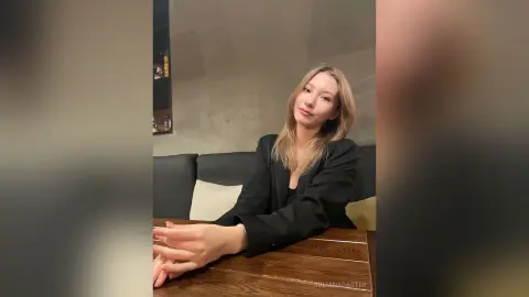 Smiling young woman in a black blazer sitting at a wooden table in a moody, dimly lit cafe setting.