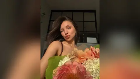 Beautiful woman in a brown top holding a large bouquet of pink peonies in a sunlit room.