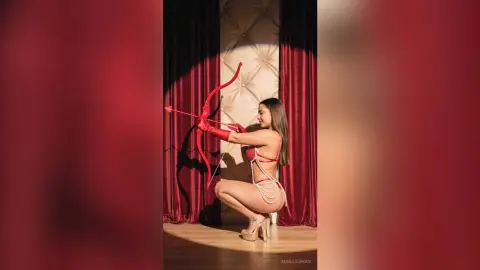 Woman in red lingerie and pearl accessories posing with a bow against red curtains in a dramatic studio setting.