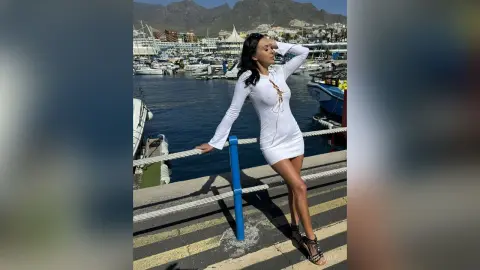 Elegant woman in a white long-sleeve mini dress posing on a sunny marina pier by the ocean.