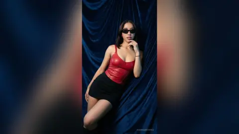 Stylish woman in a red corset and black skirt posing against blue velvet curtains for a moody fashion shoot.