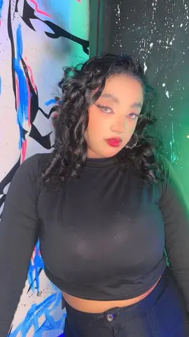 Young woman with curly hair in a black long-sleeve top posing against colorful graffiti street art.