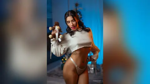 Young woman in a white long-sleeve crop top and beige briefs holding a teddy bear in a moody blue room.
