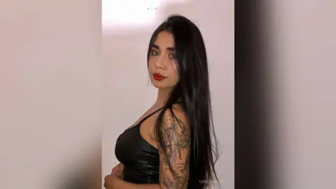 Young woman with long dark hair and red lipstick wearing a black satin camisole in a minimalist indoor setting.
