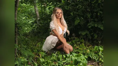 Gia poses in a white lace dress while kneeling in a lush, green forest setting.