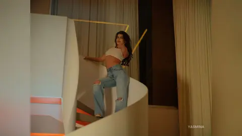 Stylish woman in a white crop top and blue jeans posing on a modern white staircase in warm lighting.